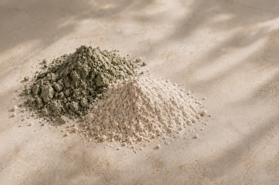 Two small piles of loose powder, one muted olive green and one light beige, arranged on a warm beige surface with soft leaf shadows.