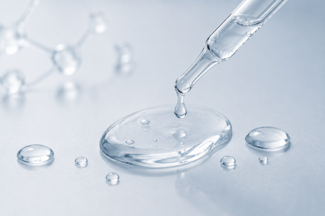 A clear glass dropper dispensing transparent liquid onto a smooth light blue surface, with several droplets and a blurred molecular model in the background.